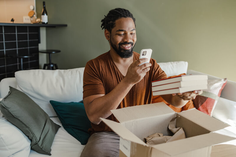 Young African American man is at home. He is unpacking the delivered package and taking a picture of the books he ordered.