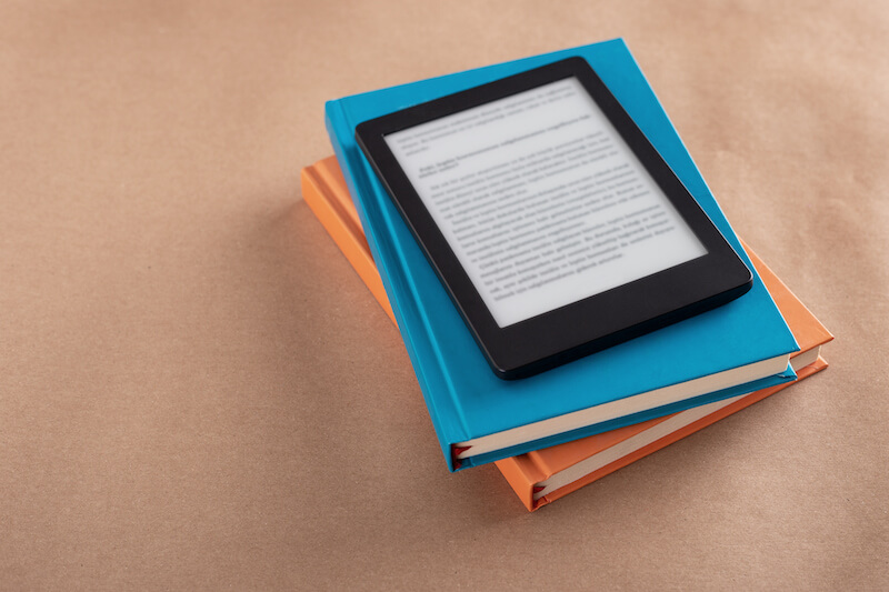 An E-book reader that is turned on placed on a stack of books.