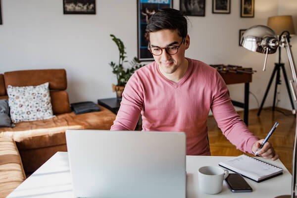 A young man with brown hair and glasses with a laptop at home, he is working and writing notes in a notebook